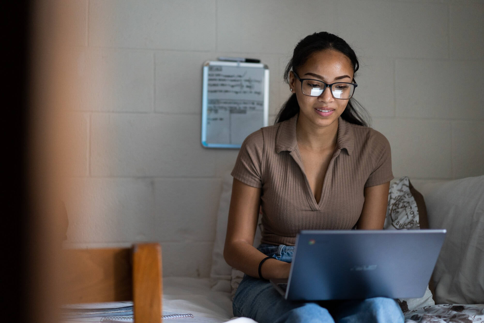 A woman with long dark hair sits on a bed in a room with cinderblock walls, wearing a short-sleeve tan collared shirt and blue jeans. She is working on a laptop and has a notebook beside her on the bed.